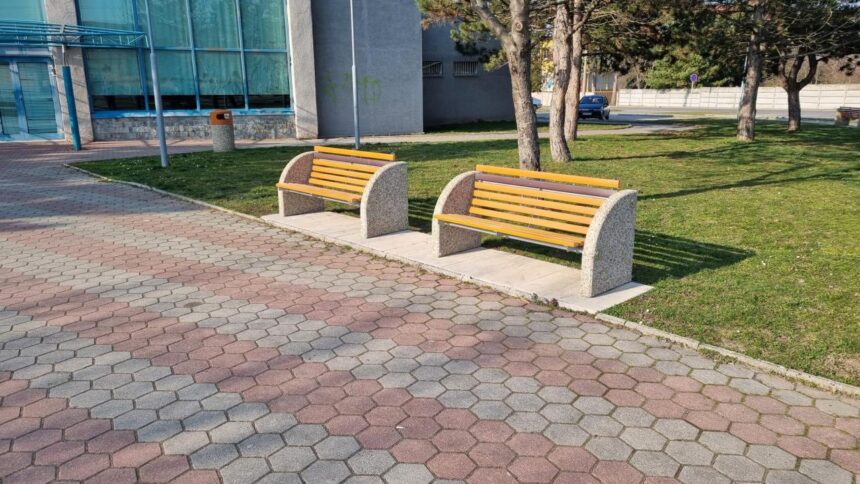 Dve nové lavičky umiestnené na chodníku pred budovou Domu kultúry v Gbeloch, v pozadí sú stromy a parkovisko.