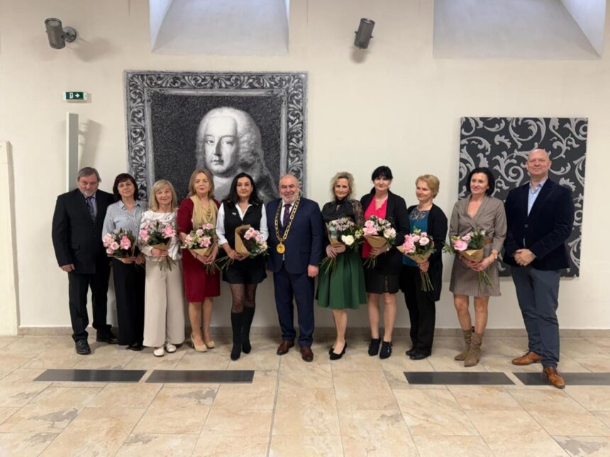 Skupina pedagógov držiacich kvety počas slávnostného stretnutia pri príležitosti Dňa učiteľov v Holíči, v pozadí obraz muža.