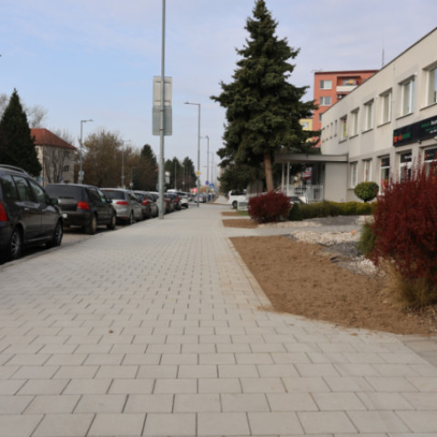Ulica v Senici s novými chodníkmi a stromami, viditeľné parkovacie miesta a zeleň pozdĺž cesty.