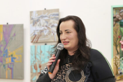 Žena hovorí do mikrofónu počas literárneho večera v Záhorskom osvetovom stredisku, v pozadí sú obrazy na stene.