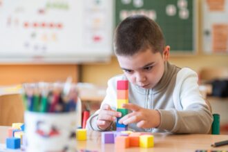 Chlapec stavia vežu z farebných kociek v školskom prostredí s tabuľou a školskými pomôckami v pozadí.