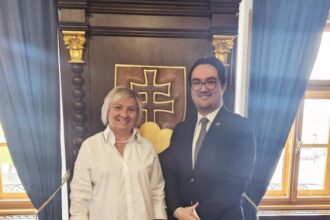 Dvaja muži stojaci pred historickým pozadím s heraldickým symbolom, jeden v bielej košeli a druhý v obleku s kravatou, usmievajú sa počas oficiálneho stretnutia v Skalici.