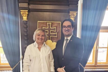 Dvaja muži stojaci pred historickým pozadím s heraldickým symbolom, jeden v bielej košeli a druhý v obleku s kravatou, usmievajú sa počas oficiálneho stretnutia v Skalici.