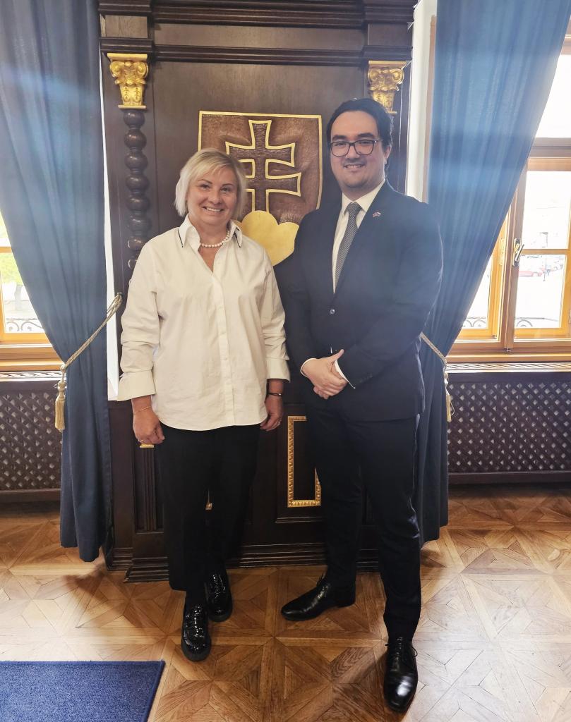 Dvaja muži stojaci pred historickým pozadím s heraldickým symbolom, jeden v bielej košeli a druhý v obleku s kravatou, usmievajú sa počas oficiálneho stretnutia v Skalici.