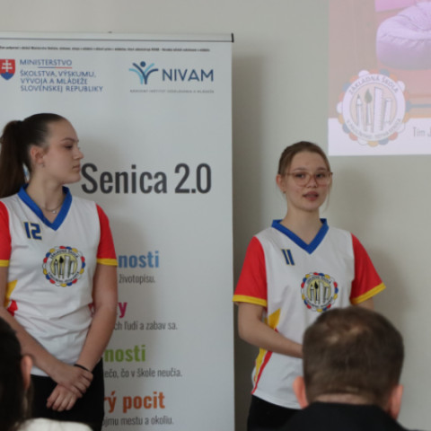 Dve dievčatá v športových dresoch prezentujú na podujatí Škola inovácií v Senici, s logom NIVAM a textom Senica 2.0 v pozadí.