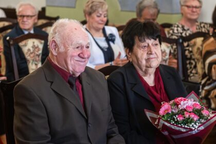 Dvojica starších ľudí sediacich na stoličkách, muž v hnedom obleku a žena v čiernom kabáte s červeným šálom drží kyticu kvetov, oslavujúca diamantovú svadbu.