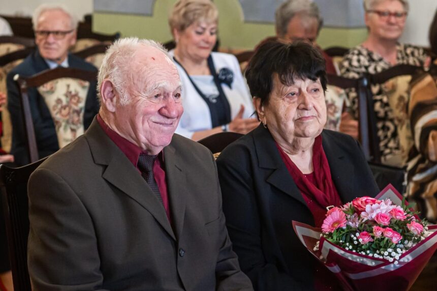 Dvojica starších ľudí sediacich na stoličkách, muž v hnedom obleku a žena v čiernom kabáte s červeným šálom drží kyticu kvetov, oslavujúca diamantovú svadbu.