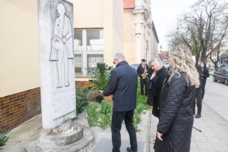 Fotografia z pietnej spomienky pri pamätníku Františka Krištofa Veselého, kde ľudia kladú kvety k pamätníku s budovou a stromami v pozadí.