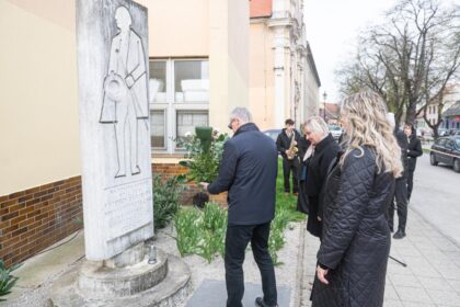 Fotografia z pietnej spomienky pri pamätníku Františka Krištofa Veselého, kde ľudia kladú kvety k pamätníku s budovou a stromami v pozadí.