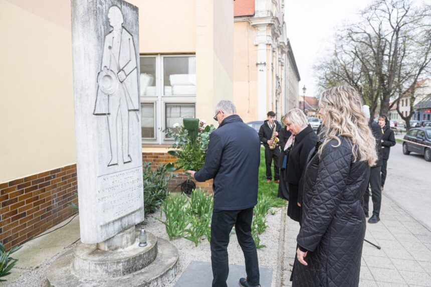 Fotografia z pietnej spomienky pri pamätníku Františka Krištofa Veselého, kde ľudia kladú kvety k pamätníku s budovou a stromami v pozadí.