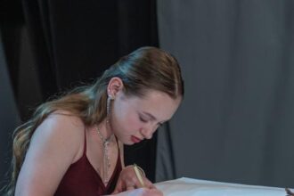 Mladá žena v červenom šate zapisuje do knihy, na stole je kvetinová dekorácia, ilustrujúca slávnostné oceňovanie športovcov v Skalici.