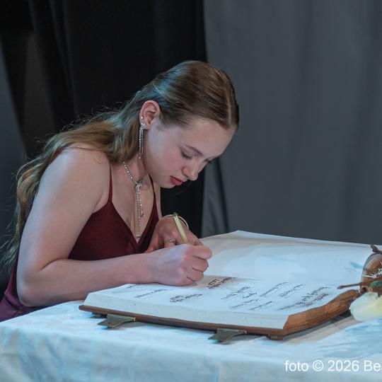 Mladá žena v červenom šate zapisuje do knihy, na stole je kvetinová dekorácia, ilustrujúca slávnostné oceňovanie športovcov v Skalici.