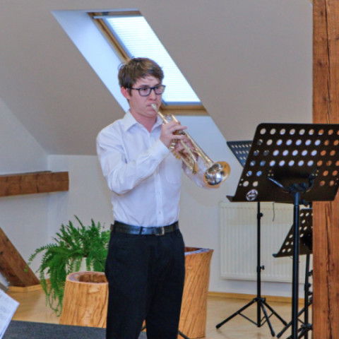 Mladý muž v bielej košeli a čiernych nohaviciach hrá na trúbke počas koncertu senických hudobníkov vo Veľkých Pavloviciach.