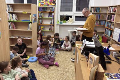Muž číta knihu deťom sediacim na podlahe v knižnici, v pozadí sú police s knihami.