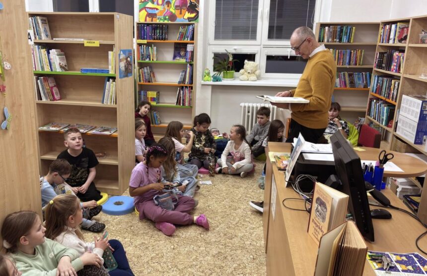 Muž číta knihu deťom sediacim na podlahe v knižnici, v pozadí sú police s knihami.