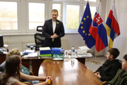 Muž v obleku sedí pri stole s niekoľkými deťmi, na stole sú dokumenty a fľaše s vodou, v pozadí sú vlajky Slovenska, Európskej únie a ďalšie.