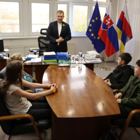 Muž v obleku sedí pri stole s niekoľkými deťmi, na stole sú dokumenty a fľaše s vodou, v pozadí sú vlajky Slovenska, Európskej únie a ďalšie.