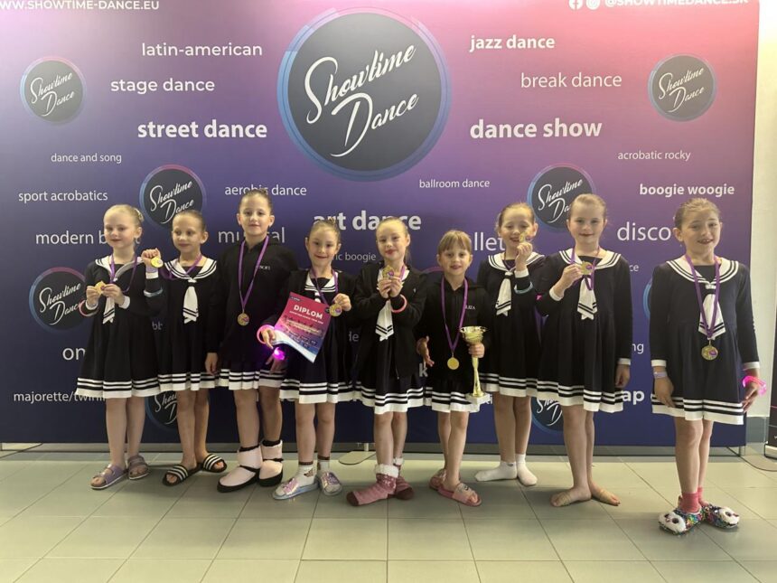 Skupina detí v čiernych kostýmoch s bielymi pruhmi, držia medaily a diplomy po tanečnej súťaži, na pozadí logo Showtime Dance.