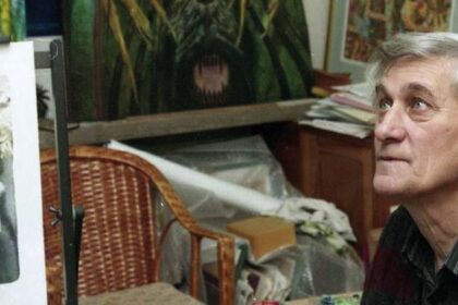 Starší muž sediaci pri stole s vystaveným obrazom, v pozadí ďalšie umelecké diela.