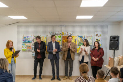 Účastníci podujatia Envirodizajn 2026 v Senici v miestnosti s výstavou detských kresieb, žena s mikrofónom prednáša, ostatní sa usmievajú a tlieskajú.