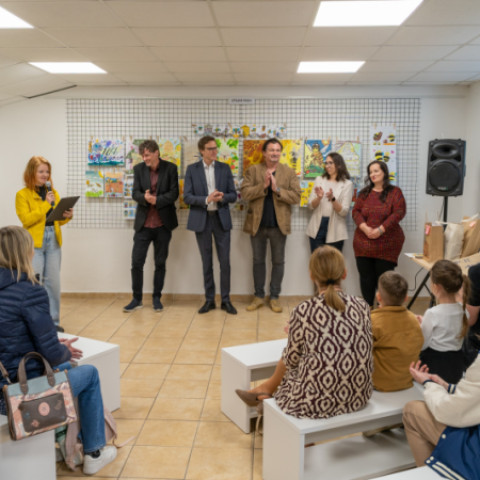 Účastníci podujatia Envirodizajn 2026 v Senici v miestnosti s výstavou detských kresieb, žena s mikrofónom prednáša, ostatní sa usmievajú a tlieskajú.