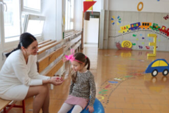 Žena sedí na lavičke a rozpráva sa s dieťaťom sediacim na modrej gymnastickej lopte v interiéri školy počas zápisu prvákov v Senici.