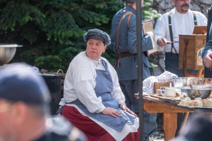 Žena v bielom oblečení a zástere sedí pri stole s jedlom, v pozadí muži v historických kostýmoch počas podujatia Varenie s c. a k. vojakmi v Skalici.