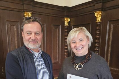 Dvaja ľudia si podávajú ruky, muž a žena, usmievajú sa, držia dokument, drevené pozadie s dekoratívnymi prvkami.