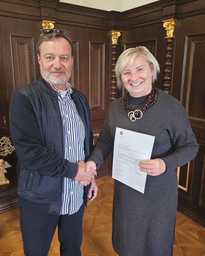 Dvaja ľudia si podávajú ruky, muž a žena, usmievajú sa, držia dokument, drevené pozadie s dekoratívnymi prvkami.