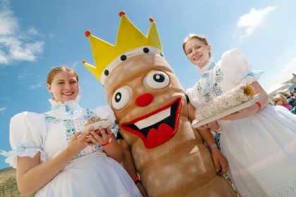 Dve dievčatá v tradičných krojoch držia tanieriky s koláčikmi, medzi nimi stojí maskot v tvare koláča s korunkou, na pozadí je jasná obloha a ľudia na festivale.