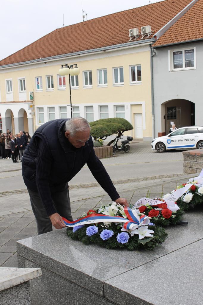 Muž kladie veniec s modrými a červenými kvetmi a páskou s nápisom MESTO na pamätník padlých vojakov na Námestí mieru v Holíči počas pietneho aktu.