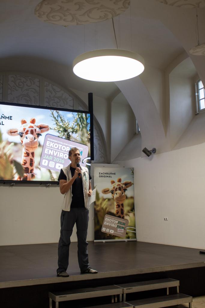 Muž na pódiu hovorí do mikrofónu počas festivalu EKOTOPFILM v Holíči, v pozadí veľké plátno s nápisom festivalu a obrázkom žirafy.