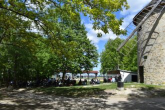 Skupina ľudí sediacich pod stromami na podujatí Majáles v areáli Veterného mlyna v Holíči, v pozadí vidieť budovu mlyna a stromy.