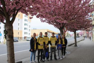 Skupina šiestich ľudí v žltých tričkách s nápisom Deň narcisov stojí na mestskej ulici s rozkvitnutými stromami v pozadí počas charitatívnej zbierky v Holíči.