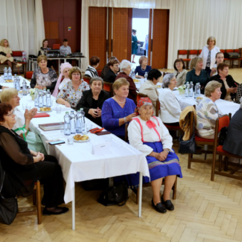 Skupina žien sediacich pri stoloch na podujatí Babička roka 2026 v Senici, niektoré sa usmievajú a tlieskajú.