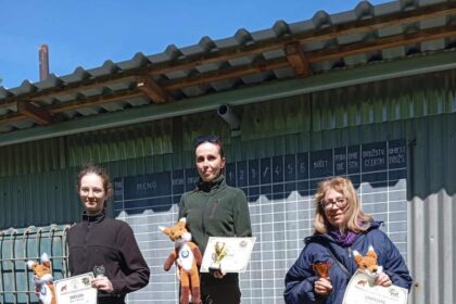 Tri osoby na stupňoch víťazov počas streleckej súťaže Holíčska Líška, držia diplomy a plyšové hračky, na pozadí je tabuľa s výsledkami.