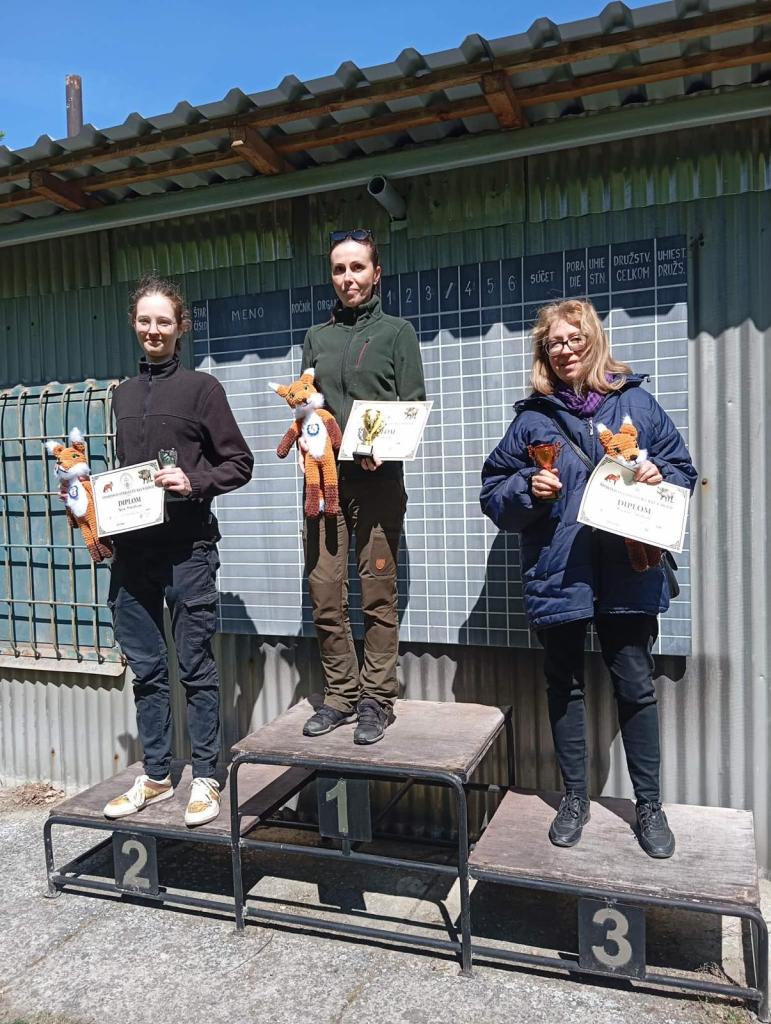 Tri osoby na stupňoch víťazov počas streleckej súťaže Holíčska Líška, držia diplomy a plyšové hračky, na pozadí je tabuľa s výsledkami.
