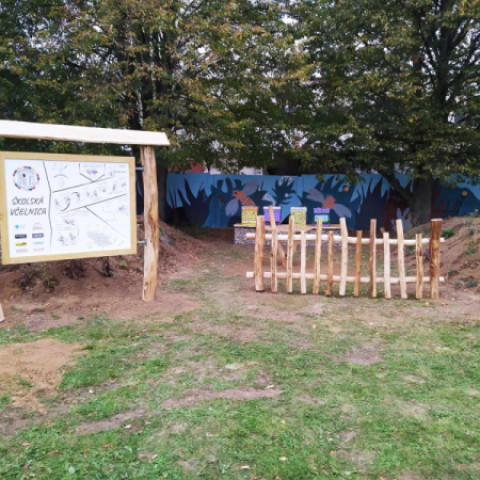 Záhrada s informačnou tabuľou a dreveným plotom, farebné maľby na stene a zeleň v pozadí, ilustrujúca komunitný projekt v Senici.