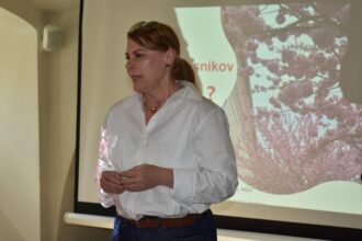Žena v bielom tričku prednáša na podujatí o skríningu prsníkov, na pozadí je premietaná prezentácia s kvetmi a textom.