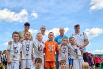Detský futbalový tím v bielych dresoch pózuje s medailami a trofejou na futbalovom štadióne v Senici počas Medzinárodného Memoriálu Fridricha Huttu.
