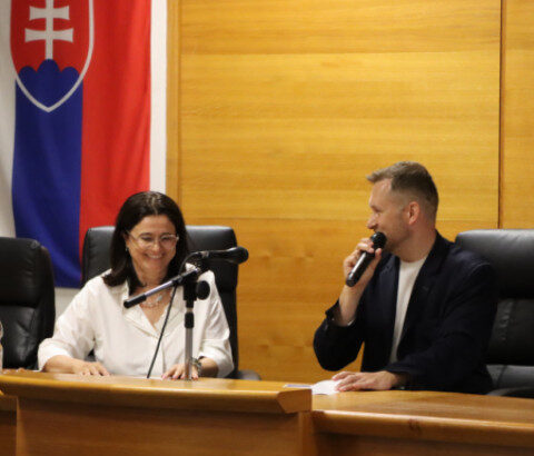 Dvaja ľudia sediaci za stolom počas diskusie na seminári o inváznych rastlinách v Senici, v pozadí slovenská vlajka.
