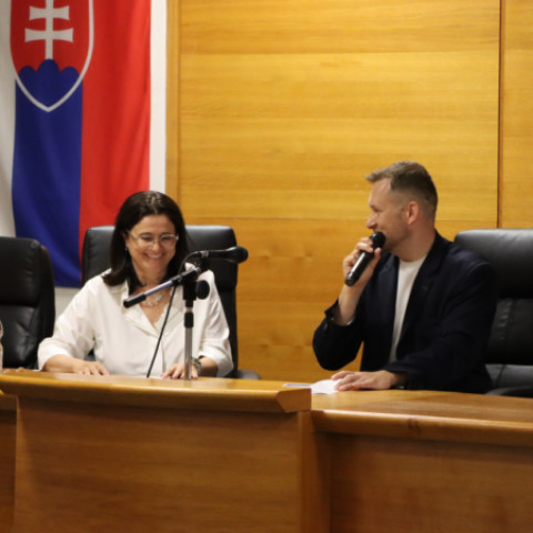 Dvaja ľudia sediaci za stolom počas diskusie na seminári o inváznych rastlinách v Senici, v pozadí slovenská vlajka.