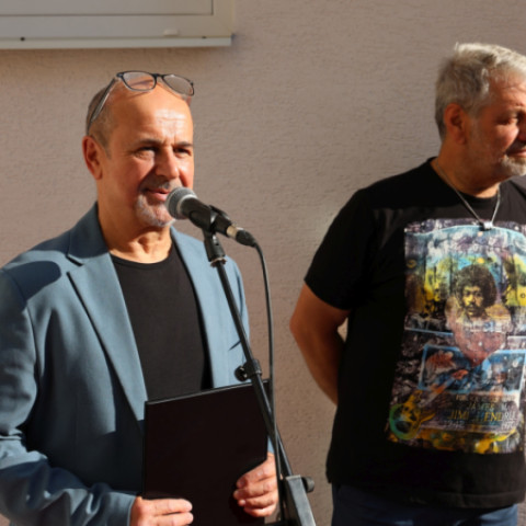 Dvaja muži v galérii počas slávnostného otvorenia, jeden hovorí do mikrofónu, druhý stojí vedľa neho.
