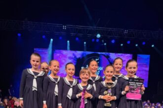 Skupina mladých balerínok v tmavomodrých kostýmoch s bielymi detailmi stojí na pódiu, držia trofej a ocenenie. Dievčatá sú bosé a usmievajú sa, na pozadí je nápis "Showtime 2 Dance".
