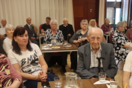 Skupina starších ľudí sedí pri stole s pohármi vody a taniermi s koláčikmi v tanečnej sále Domu kultúry v Senici počas slávnostného stretnutia seniorov oslavujúcich životné jubileá.