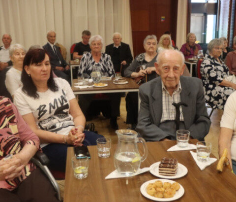 Skupina starších ľudí sedí pri stole s pohármi vody a taniermi s koláčikmi v tanečnej sále Domu kultúry v Senici počas slávnostného stretnutia seniorov oslavujúcich životné jubileá.