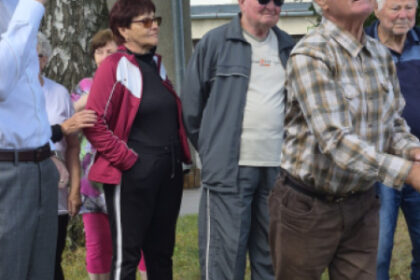 Skupina starších ľudí vonku počas športových hier v Senici, niektorí sa aktívne zapájajú do športových aktivít, iní sledujú dianie.