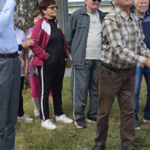 Skupina starších ľudí vonku počas športových hier v Senici, niektorí sa aktívne zapájajú do športových aktivít, iní sledujú dianie.