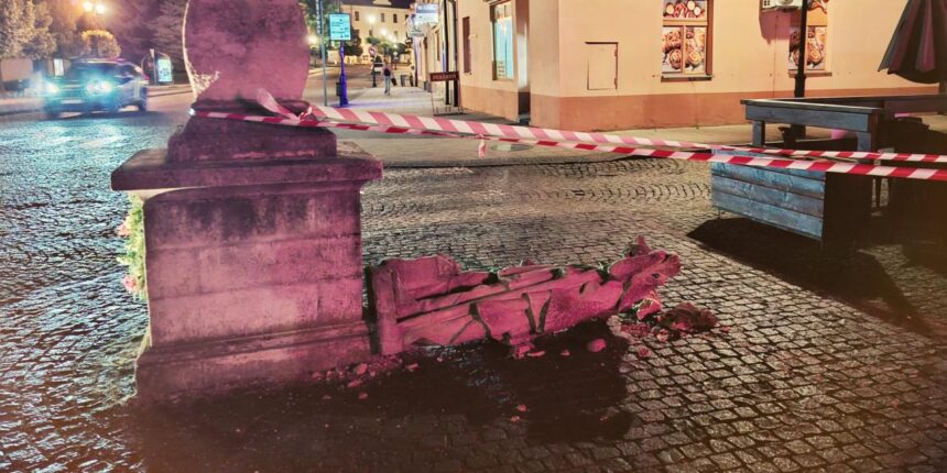 Zničená socha sv. Floriána ležiaca na zemi v centre Skalice, obklopená červenobielym pásmom, v pozadí vidieť budovy a pouličné osvetlenie.