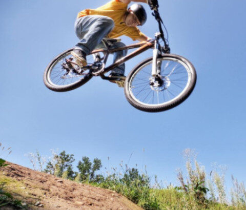 Chlapec na bicykli skáče cez prekážku na modrom pozadí s oblohou a zeleňou, ilustrujúci bikepark pre mladých v Senici.